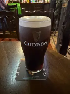 A Guinness pint glass with a creamy head rests on a branded coaster atop a dark wooden table in a dimly lit pub, with the bar and chairs softly blurred in the background.
