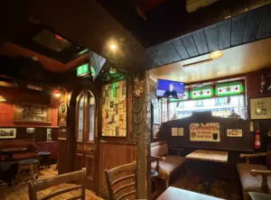 Cozy, dimly lit pub interior with wooden furniture, vintage decor, stained glass, and a Guinness pint glass sign. TV screens show a man talking. Seating booths and walls display various memorabilia and mementos.