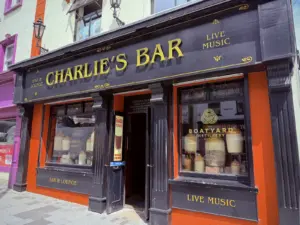 Charlie's Bar exterior features black and gold signage, a Guinness pint glass, and a Boatyard Distillery sign in the window. With bright orange trim, the pub welcomes guests through an open doorway beside whiskey barrels.