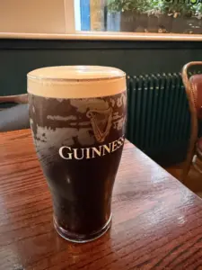 A pint glass of Guinness stout with a creamy head sits on a wooden table in a cozy pub setting, with a chair and green radiator visible in the background.