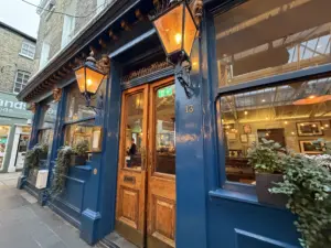 A view of a traditional pub exterior with dark blue walls, large wooden double doors, glowing lanterns, and potted plants. The number 13 is above the door, and the warmly lit interior is visible through the windows.