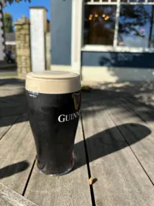 A pint of Guinness stout sits on a wooden outdoor table in sunlight, casting a shadow. A blue building and window are in the background.