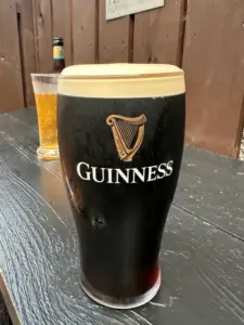 A pint of Guinness with a creamy head sits on a wooden table outdoors, with a glass of pale beer and a bottled drink blurred in the background.