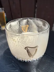 A Guinness glass with remnants of beer foam sits on a dark wooden table outdoors, with another beverage glass in the background and sunlight casting shadows.