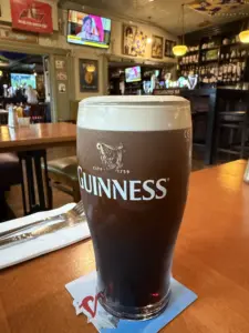 A pint of Guinness stout sits on a bar table in a cozy pub, with utensils on the left, shelves of bottles in the background, and a TV showing a woman’s face above the bar.