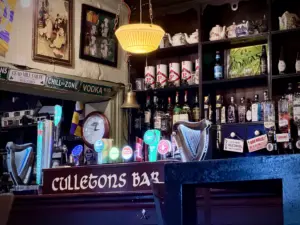 A cozy bar interior with a wooden counter labeled "Culleton's Bar," various beer taps, bottles of liquor on shelves, framed photos, and a yellow hanging lamp creating a warm, inviting atmosphere.