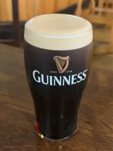 A Guinness pint glass with a creamy head sits on a wooden table, featuring the iconic logo and harp emblem. The background reveals a softly blurred pub interior.