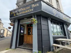 Outside view of Celtic Marine Bar & Restaurant, featuring a black and gold sign with a Guinness pint glass, hanging flower baskets, wooden doors, large windows, and outdoor wooden benches on a corner street.