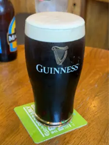 A pint of Guinness stout with a thick creamy head, served in a branded Guinness pint glass on a green coaster, sits on a wooden table. Another bottle is partially visible in the background, evoking classic pub vibes.
