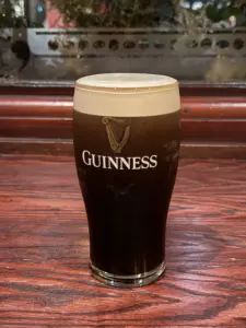 A Guinness pint glass with a creamy, tan head sits on a wooden table in a pub setting. The logo and harp symbol are visible on the glass, with blurred greenery and wood paneling in the background.