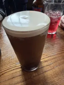 A pint glass filled with dark beer, topped with a thick, creamy head, sits on a wooden table. Nearby are a glass with a red drink and a brown bottle, with hands and blurred figures in the background.