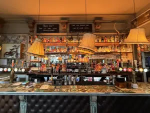 A cozy bar with a patterned countertop, vintage hanging lamps, shelves filled with bottles, and chalkboard menus on the wall behind the counter. The decor features eclectic and antique touches.