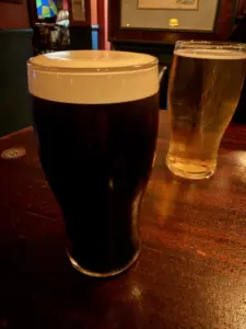 A pint of dark stout with a creamy head and a pint of light-colored beer sit on a wooden pub table, with a dimly lit bar interior in the background.