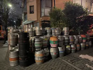 A large stack of metal beer kegs, some painted in colorful stripes, is arranged on a sidewalk at night near a brick building with plants and a tree visible in the background.
