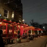 A cozy pub called "The Dog & Bell" glows with warm red lighting at night. Outdoor seating under red canopies is occupied, and string lights hang above; a person walks by on the dimly lit street.