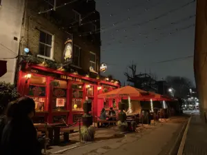 A cozy pub called "The Dog & Bell" glows with warm red lighting at night. Outdoor seating under red canopies is occupied, and string lights hang above; a person walks by on the dimly lit street.