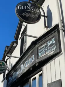 A pub sign reading "The Blue Devon" with another sign below stating "We trade in thousands of beers, mugs, gossip." Above is a circular Guinness sign. The building is white with black trim under a clear blue sky.
