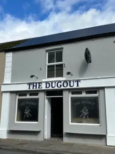 A grey and white building with a sign reading "THE DUGOUT" above the door. Two large front windows display the same logo and name. The sky is partly cloudy above the building.