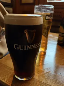 A Guinness pint glass with a creamy head sits on a wooden table beside a pint of Bulmers cider in a dimly lit pub.