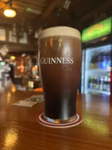 A Guinness pint glass with a creamy head sits on a coaster on a wooden bar in a pub, with blurred shelves and a refrigerator in the background.