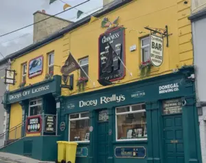 A yellow and green pub with signs reading "Dicey Reilly’s," "Dicey’s Off Licence," and featuring a Guinness pint glass. Flags and flower boxes decorate the exterior, with a yellow bin on the sidewalk outside.