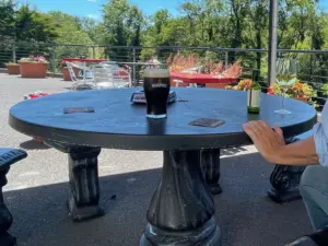 A round black outdoor table with a pint of stout, a glass of white wine, and coasters. A person’s hand rests on the table. Bright patio with flowers, trees, and lounge chairs in the background under a blue sky.