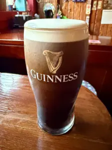 A guinness pint glass with a creamy head sits on a wooden table in a cozy pub setting, featuring the Guinness logo and harp symbol on the glass.