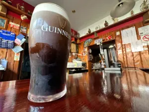 A Guinness pint glass rests on a polished wooden bar counter in a cozy pub, with taps, shelves, and various decorations visible in the background.