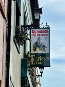 A hanging pub sign reads "Guinness for Strength" above an image of a man pulling a cart with a horse inside. Below, it says "Earley's Bar," with a Guinness pint glass featured, attached to a building with flower boxes.