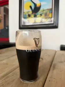 A Guinness pint glass sits partially full on a wooden table at a pub, foam circling the rim. A colorful painting is blurred in the background on a white wall.