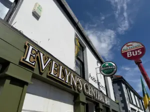 Close-up of Evelyn's Central Bar sign on a white building, featuring a Guinness pint glass pub sign and a red Bus Éireann bus stop sign against a blue sky with some clouds.