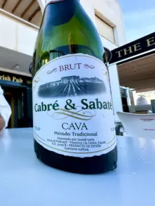 A close-up of a bottle of Cabré & Sabaté Brut Cava on an outdoor table, with a Guinness pint glass nearby and part of a pub sign visible in the background. The wine label details are clearly readable.