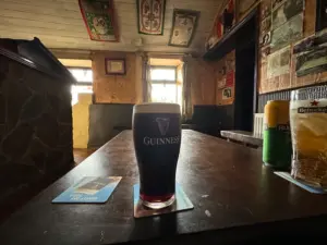A Guinness pint glass rests on a wooden table inside a cozy, dimly lit pub adorned with posters and memorabilia. Daylight streams through two small windows, casting a warm glow over the inviting scene.