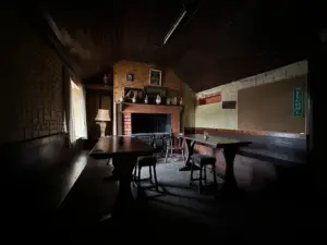 A dimly lit pub room with wooden tables and chairs, a Guinness pint glass on the table, a brick fireplace, framed pictures above the mantel, and a lamp near a curtained window. A “Welcome” sign hangs on the wall to the right.