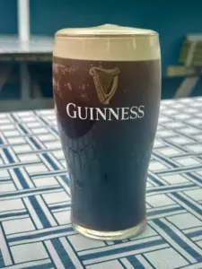 A pint of Guinness stout with a creamy head sits on a patterned blue and white tablecloth, with blurred benches and a dark wall in the background.