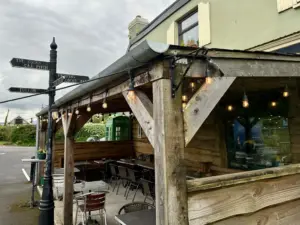 A rustic outdoor seating area of a café or pub with wooden beams, string lights, and empty tables and chairs. A vintage signpost, a green phone booth, and a Guinness pint glass are visible under a cloudy sky.