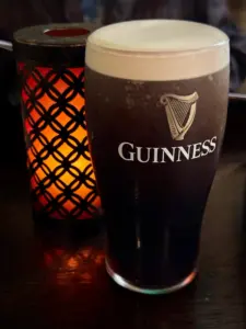 A Guinness pint glass with a creamy head sits on a dark wooden table in a cozy pub, beside a decorative lantern emitting a warm orange glow.
