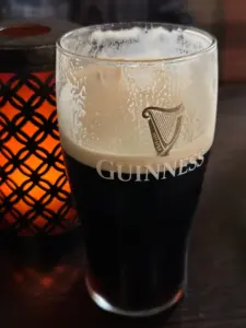 A Guinness pint glass sits on a dark wooden table beside a lit lantern with a black lattice pattern, creating a warm, cozy pub atmosphere.