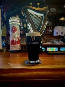 A Guinness pint glass sits on a wooden bar, with a large Guinness harp sign and a Smithwick's tap in the background, capturing the cozy atmosphere of a dimly lit pub.