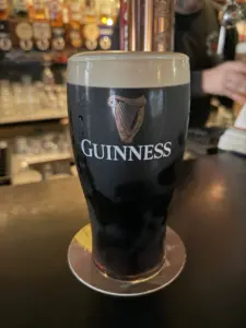 A Guinness pint glass with a creamy head sits on a coaster at a pub counter, with bar taps and glasses visible in the blurred background.