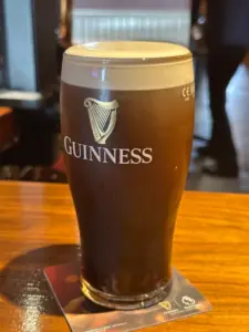 A pint glass of Guinness stout with a creamy head sits on a coaster atop a wooden table in a pub, with blurred figures and background visible.