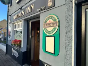 Exterior of Fordes Inn lounge with a Heineken beer sign on the gray wall, red flowers in a window box, and sunlight casting shadows on the entrance.