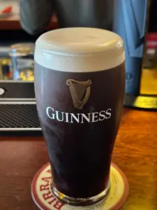 A Guinness pint glass with a creamy head sits on a coaster atop a wooden bar counter in a cozy pub, with bar equipment and a blurred background visible.