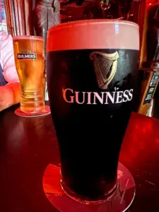 A Guinness pint glass and a glass of Bulmers cider rest on coasters atop a dark bar table in a dimly lit pub with red lighting.
