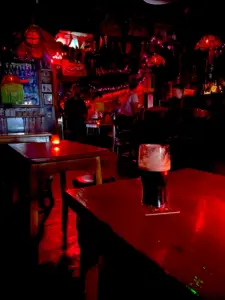 A dimly lit pub with red lighting, wooden tables, and a Guinness pint glass in the foreground. Several people sit and stand in the background, creating a cozy, intimate atmosphere.