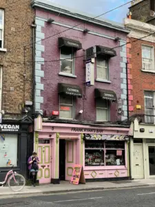A person stands outside Frank Ryan’s Bar, a purple and pink pub with Guinness signs and a pint glass logo, on a street lined with brick buildings. A pink bicycle is parked nearby, and colorful signs are visible in the window.