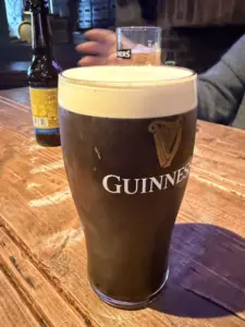 A Guinness pint glass with a creamy head rests on a rustic wooden table in a pub, with another bottle and a person’s arm visible in the background.