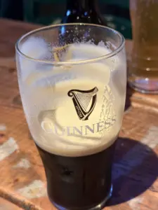 A partially finished Guinness pint glass with foam residue sits on a wooden table in a pub, with another glass and a bottle in the blurred background.