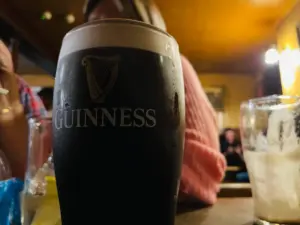 A Guinness pint glass sits on a table in a pub, with a blurred person in a pink sweater and other glasses in the background. The focus is on the rich, dark liquid with its creamy head.