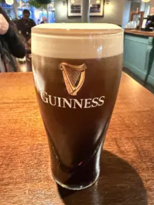 A pint of Guinness stout with a creamy head sits on a wooden table in a warmly lit pub, with blurred people and bar area in the background.